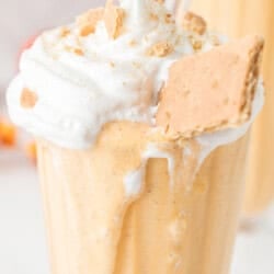 A glass filled with a creamy orange milkshake, topped with whipped cream, crushed crumbs, and a piece of graham cracker, with a straw inserted.