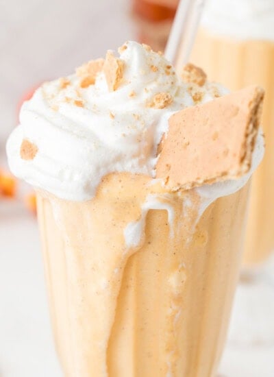 A glass filled with a creamy orange milkshake, topped with whipped cream, crushed crumbs, and a piece of graham cracker, with a straw inserted.