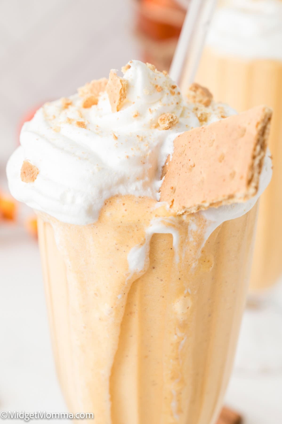 A glass filled with a creamy orange milkshake, topped with whipped cream, crushed crumbs, and a piece of graham cracker, with a straw inserted.