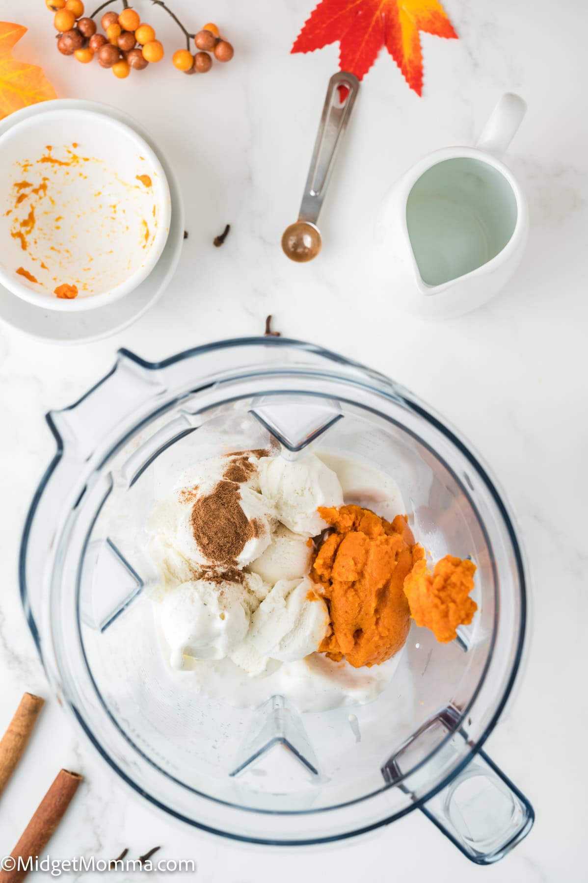 A blender contains cream cheese, pumpkin puree, and spices, surrounded by a bowl, measuring spoon, ceramic pitcher, cinnamon sticks, and fall decorations on a white surface.