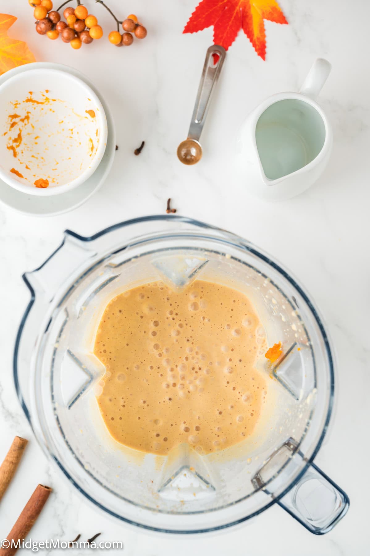A blender filled with a frothy orange mixture sits on a white countertop, surrounded by a measuring spoon, small pitcher, empty bowl, cinnamon sticks, and autumn decorations.