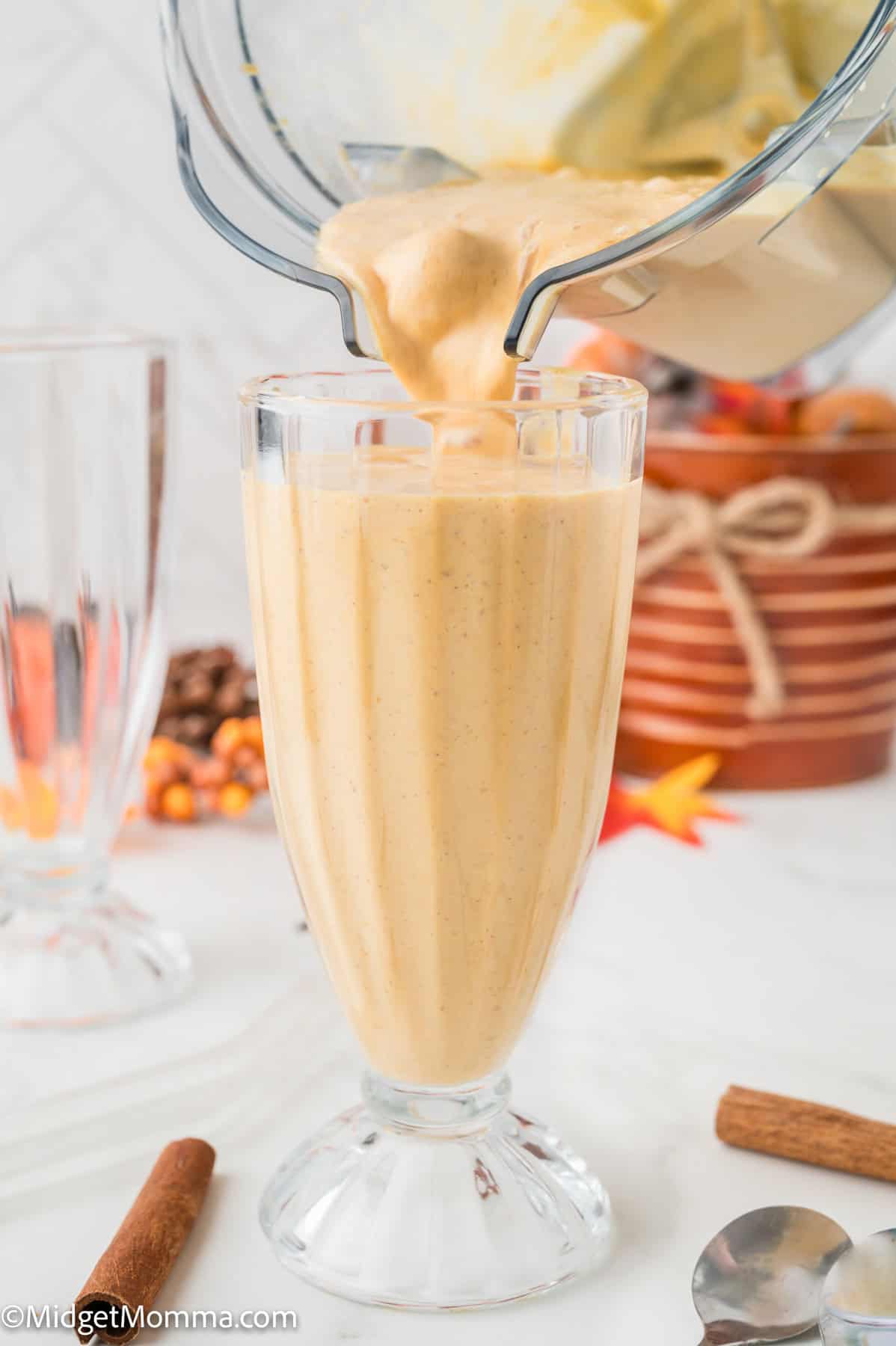 A thick pumpkin smoothie is being poured from a blender into a tall glass, with fall-themed decorations and cinnamon sticks in the background.