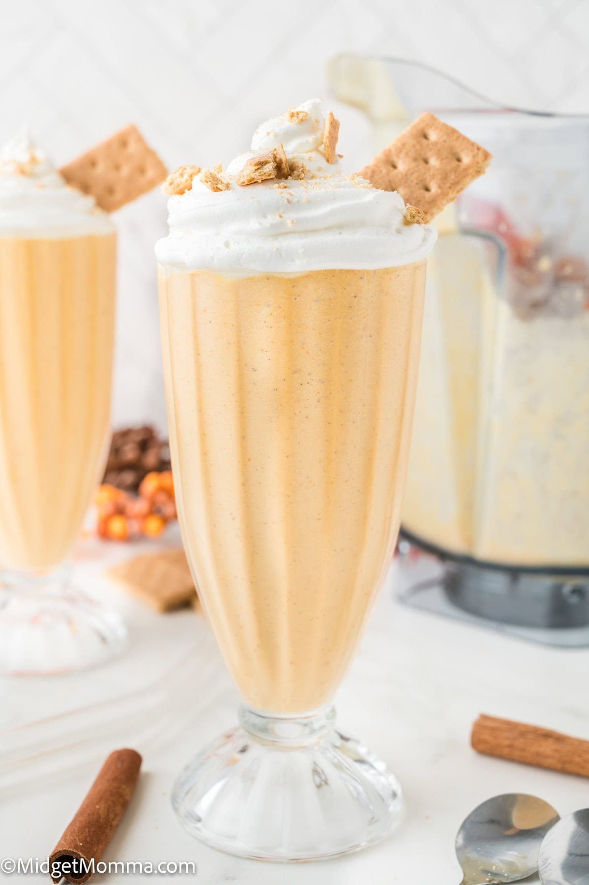 A tall glass of pumpkin milkshake topped with whipped cream, crushed graham crackers, and a graham cracker piece, with a blender and another milkshake in the background.