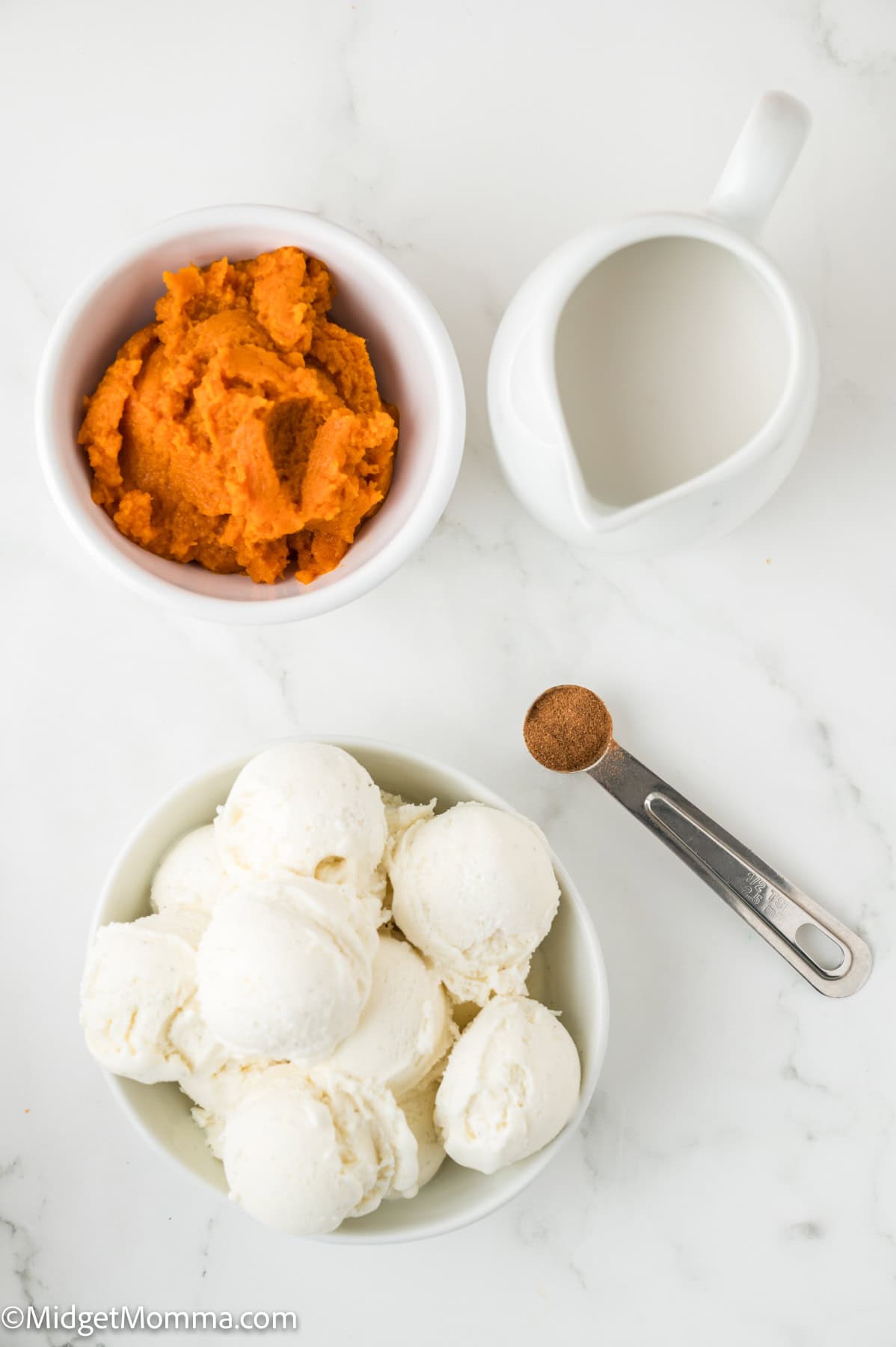 Bowls of vanilla ice cream and pumpkin puree next to a small pitcher of cream and a teaspoon of cinnamon on a white surface.