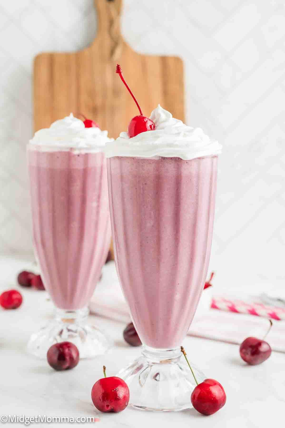 Two tall glasses of cherry milkshakes topped with whipped cream and a cherry, with fresh cherries scattered around on a white surface.