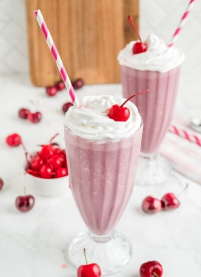 Two tall glasses of pink milkshake topped with whipped cream and a cherry, each with a striped straw. Fresh cherries are scattered around the glasses.