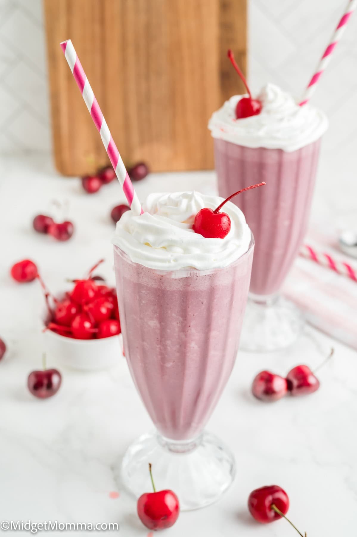 Two tall glasses of pink milkshake topped with whipped cream and a cherry, each with a striped straw. Fresh cherries are scattered around the glasses.