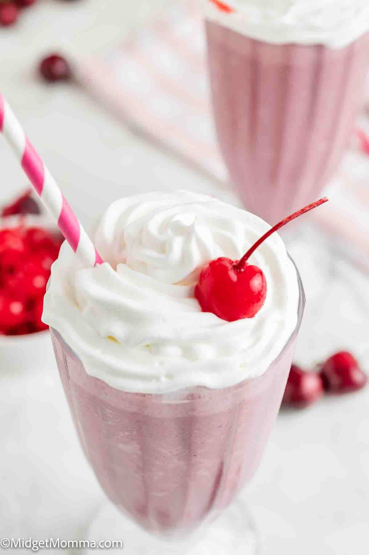 A pink milkshake topped with whipped cream and a maraschino cherry, served in a glass with a striped straw.