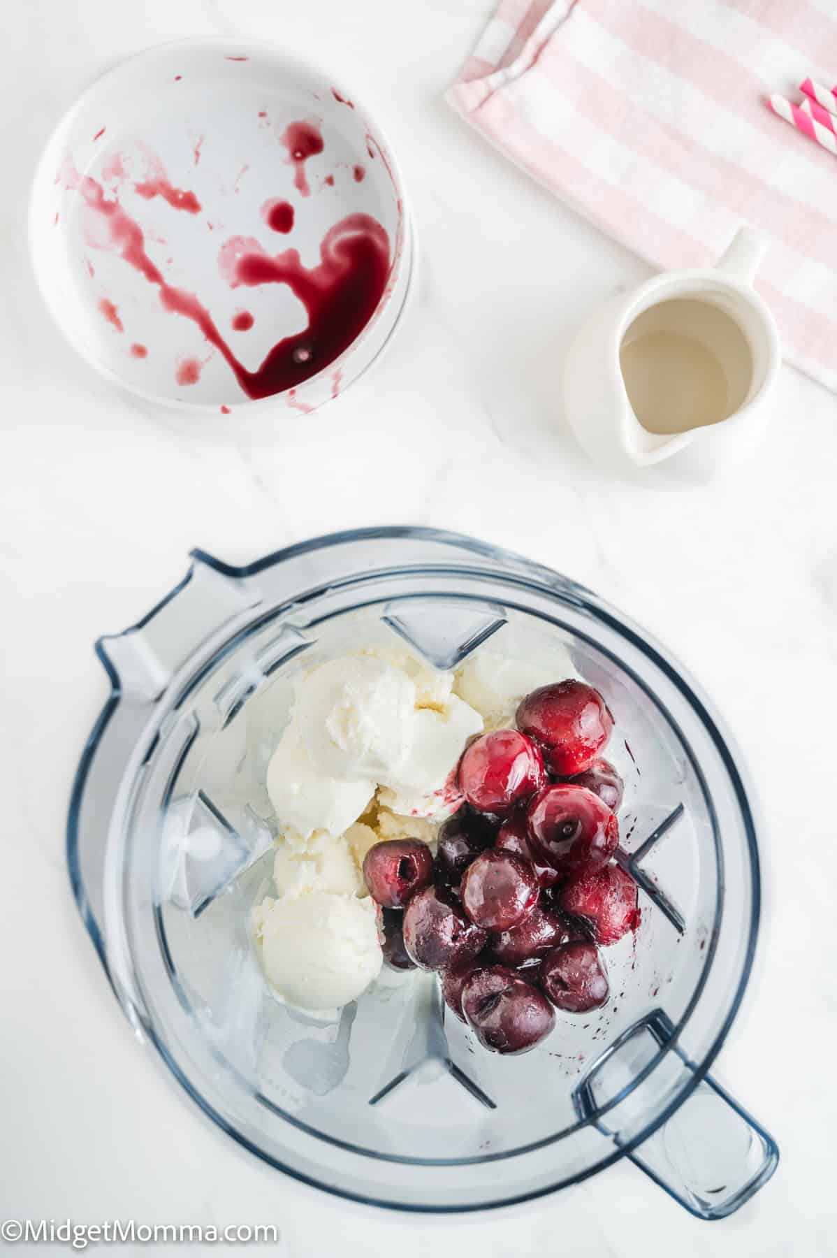 A blender bowl contains scoops of vanilla ice cream and frozen cherries. Nearby are a bowl with cherry juice stains, a striped towel, and a small white pitcher.