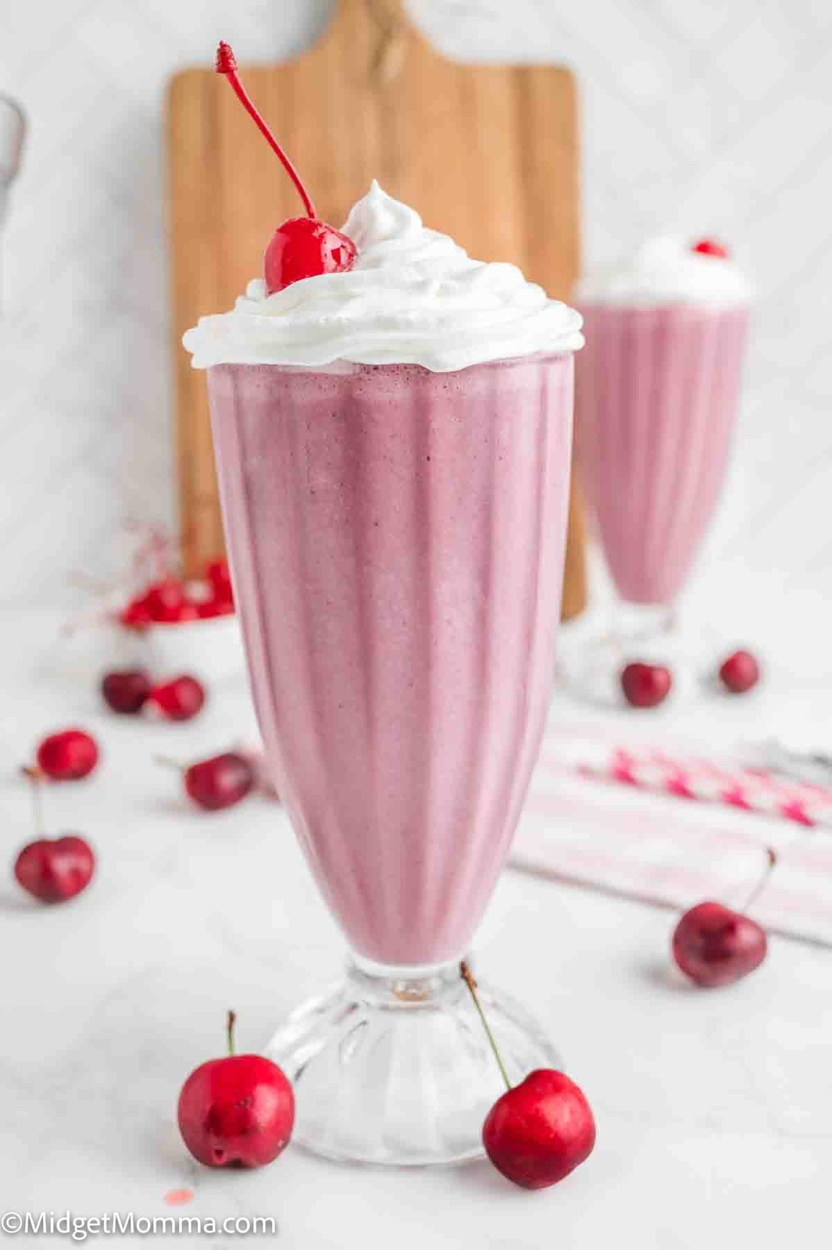A tall glass of pink milkshake topped with whipped cream and a cherry, surrounded by fresh cherries on a white surface.