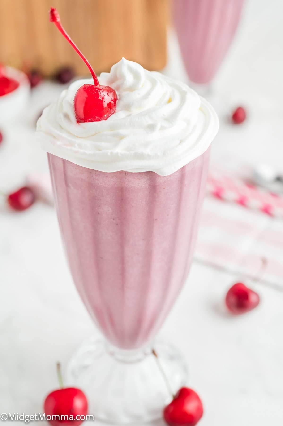 A pink milkshake in a tall glass topped with whipped cream and a cherry, with fresh cherries scattered around on a white surface.