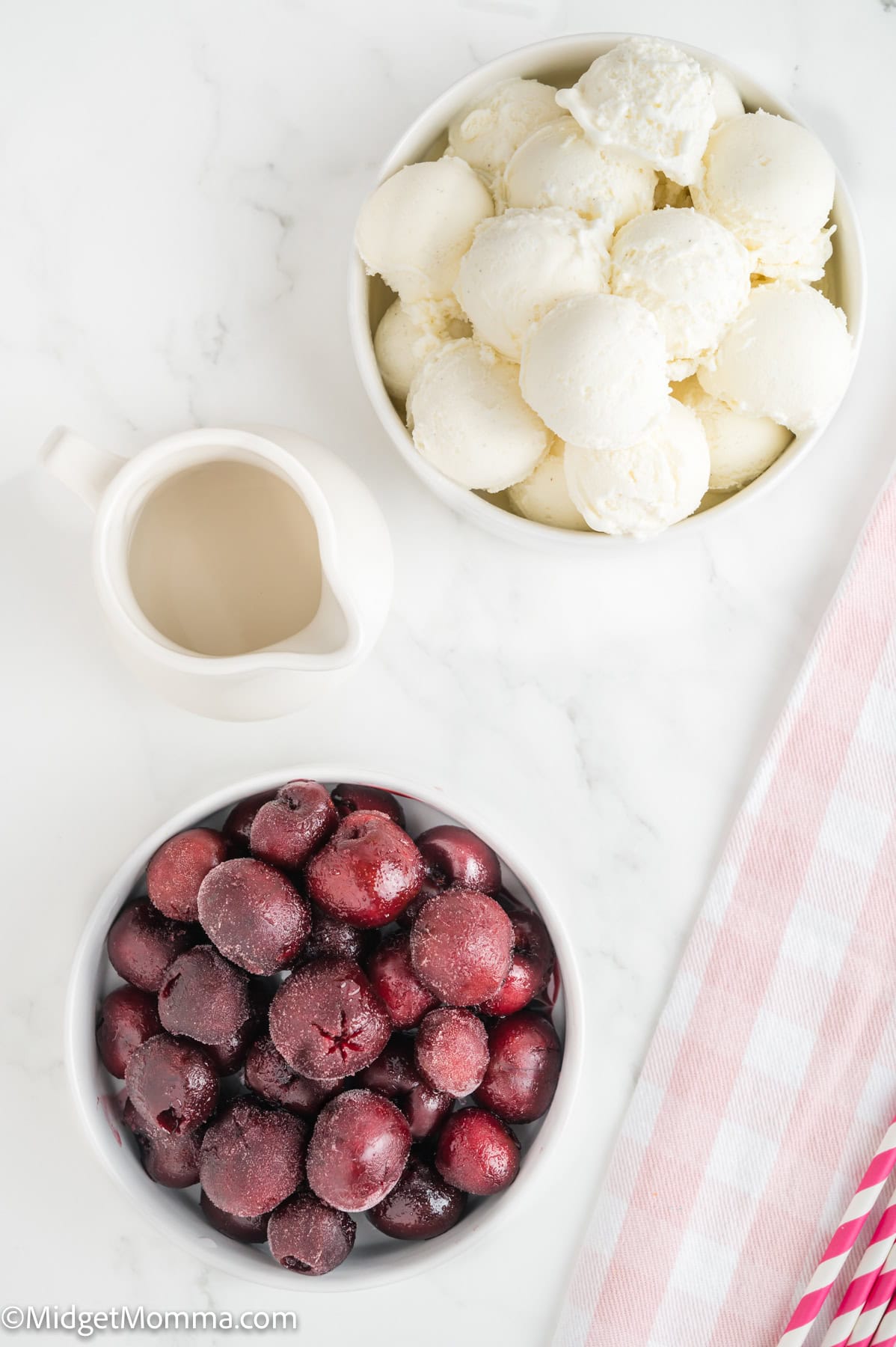 Two bowls on a white surface: one filled with vanilla ice cream scoops, the other with frozen cherries. A small pitcher and striped napkin are also visible.
