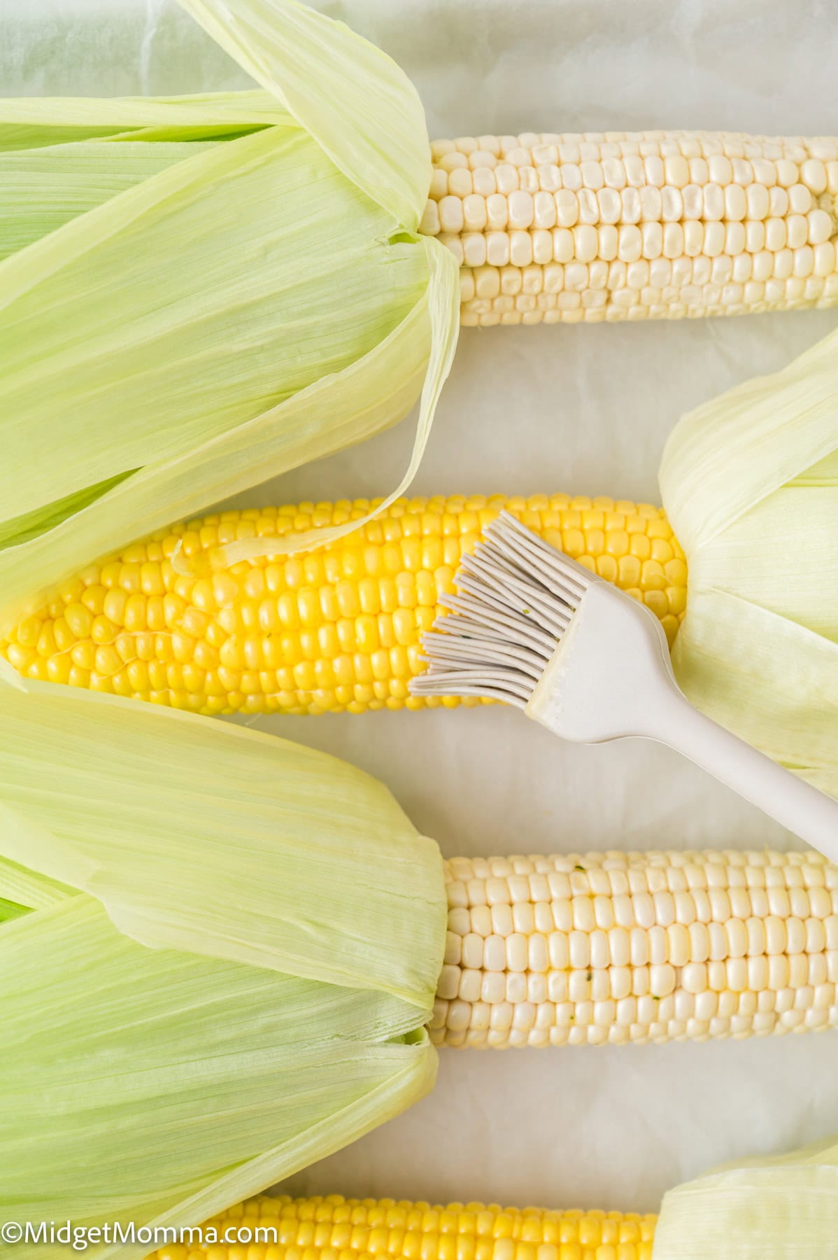 Three ears of corn, two white and one yellow, partially husked and arranged on a surface, with a basting brush resting on the yellow corn.