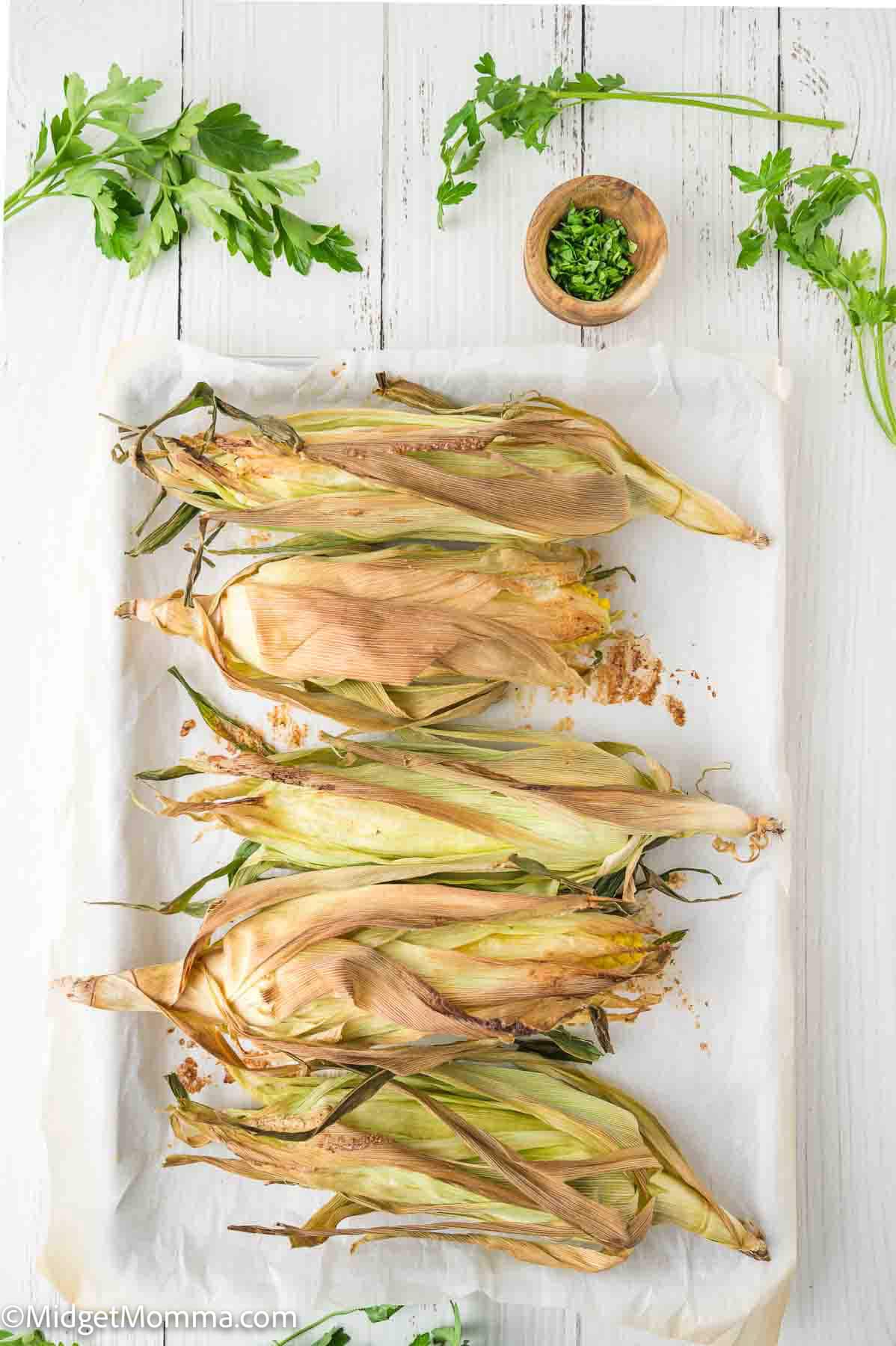 Five ears of corn with husks partially peeled back are arranged on parchment paper atop a white wooden surface, with fresh herbs and a small bowl of chopped chives nearby.
