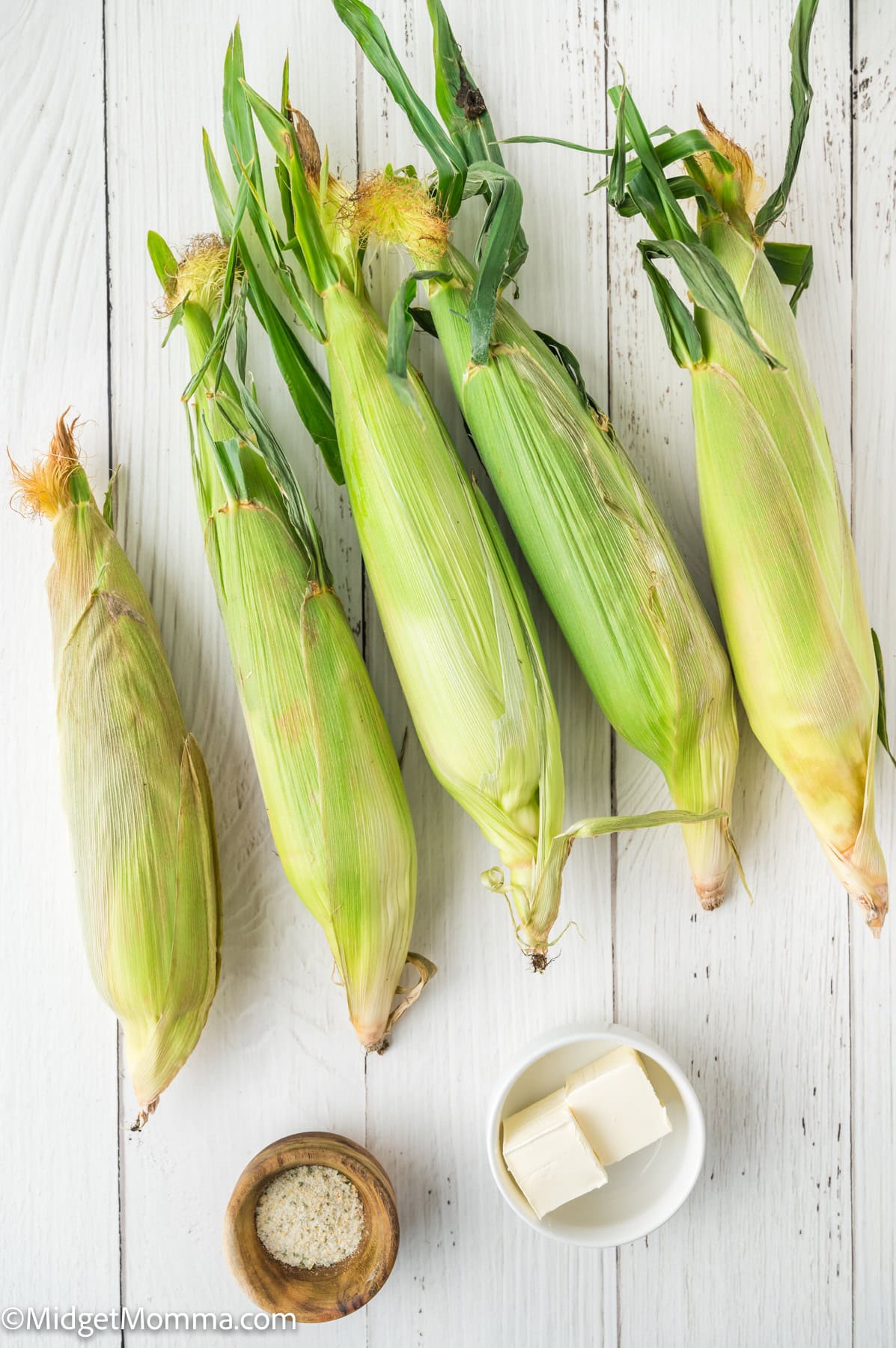 Five ears of corn in husks are arranged on a white wooden surface next to a small bowl of salt and a dish with three pieces of butter.