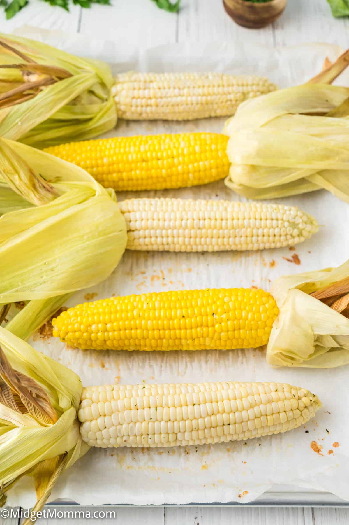 Six ears of corn, some yellow and some white, partially husked and arranged on a parchment-lined surface.