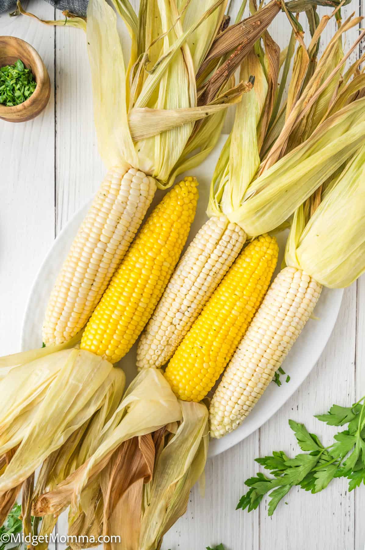 Four ears of corn with husks partially on, arranged on a white plate; two ears are yellow and two are white. Parsley and chopped herbs are nearby.