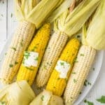 Five ears of corn with husks partially peeled back on a white plate, topped with butter and chopped herbs.