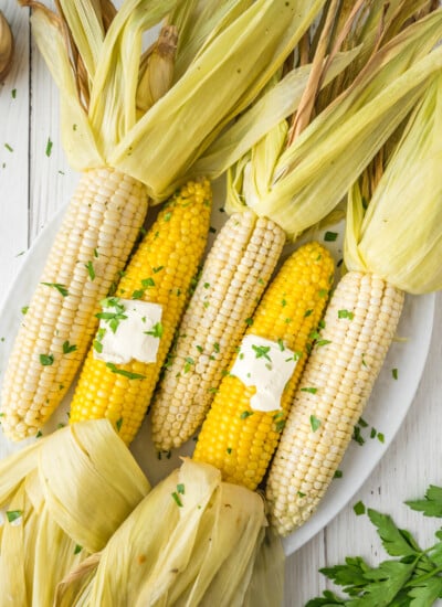 Five ears of corn with husks partially peeled back on a white plate, topped with butter and chopped herbs.