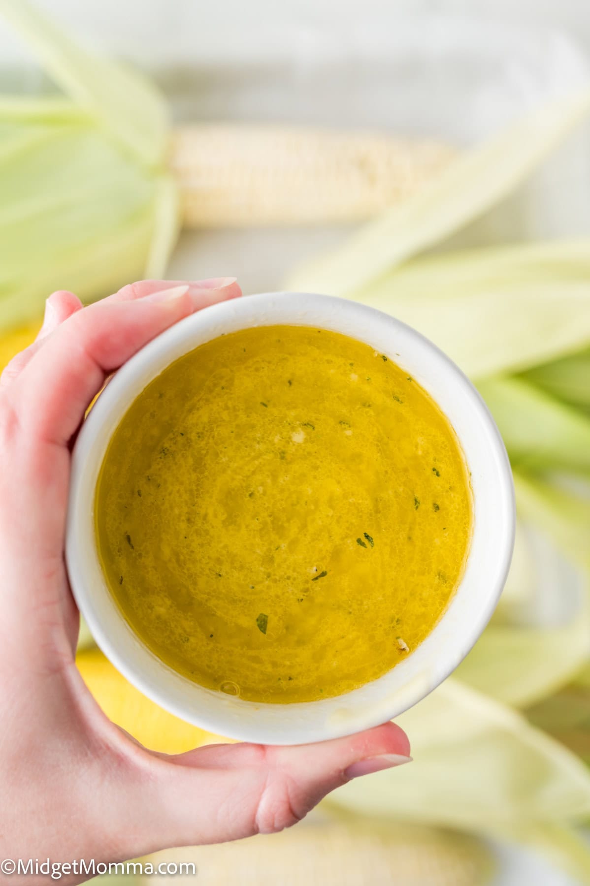 A hand holding a white bowl filled with a yellow herb butter sauce, with corn and husks visible in the background.