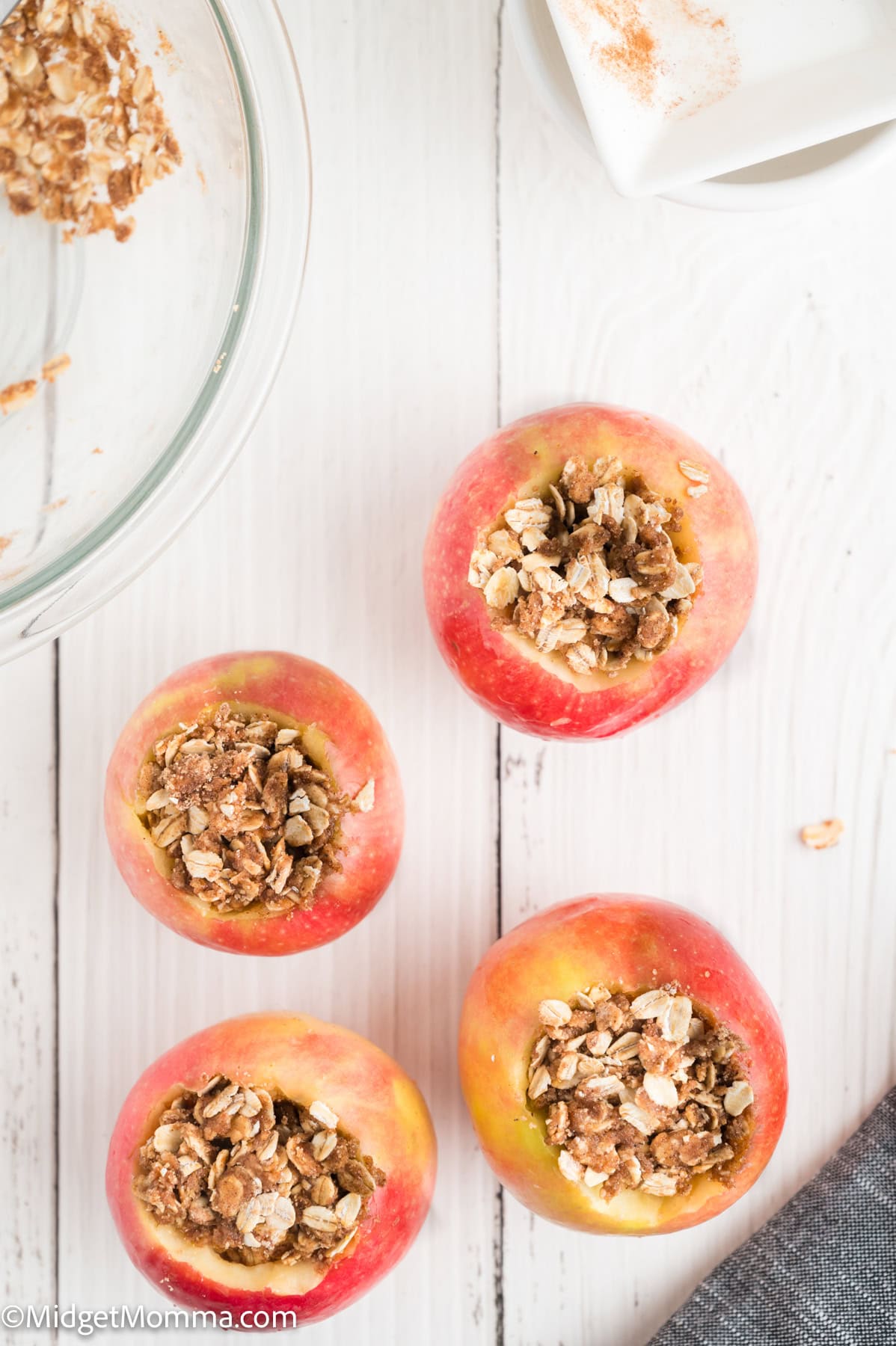 Four apples with their tops removed, filled with an oat and cinnamon mixture, are arranged on a white wooden surface next to a glass bowl and a napkin.