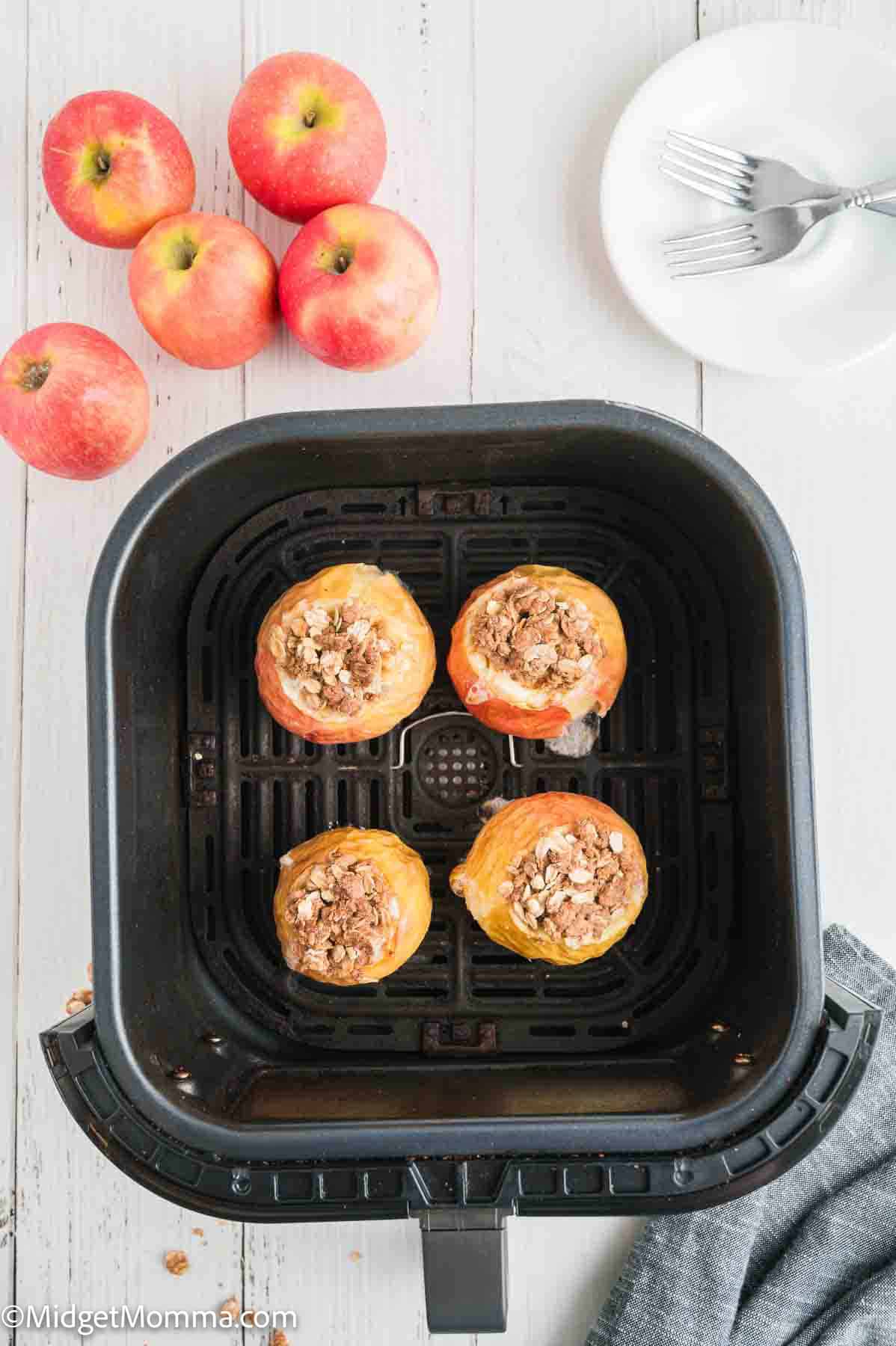 Four baked apples filled with crumble sit in an air fryer basket, with whole apples, a plate, and forks on a white wooden surface nearby.