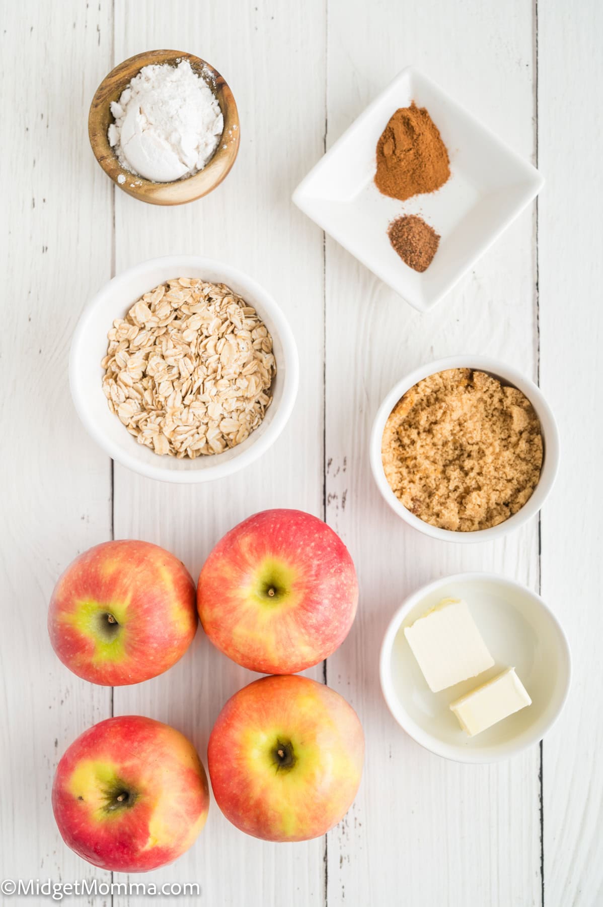 Flat lay of ingredients on a white surface: four apples, oats, brown sugar, butter, cornstarch, ground cinnamon, and nutmeg.