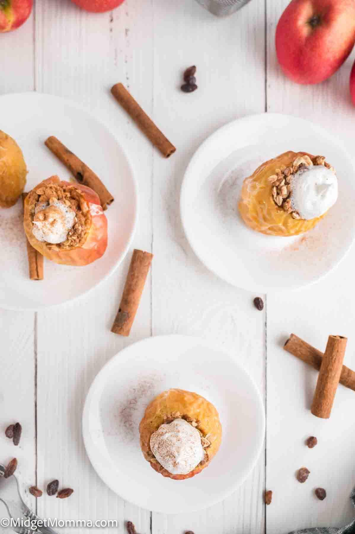 Three baked apples topped with granola and whipped cream are served on white plates, surrounded by cinnamon sticks and whole apples on a white wooden surface.
