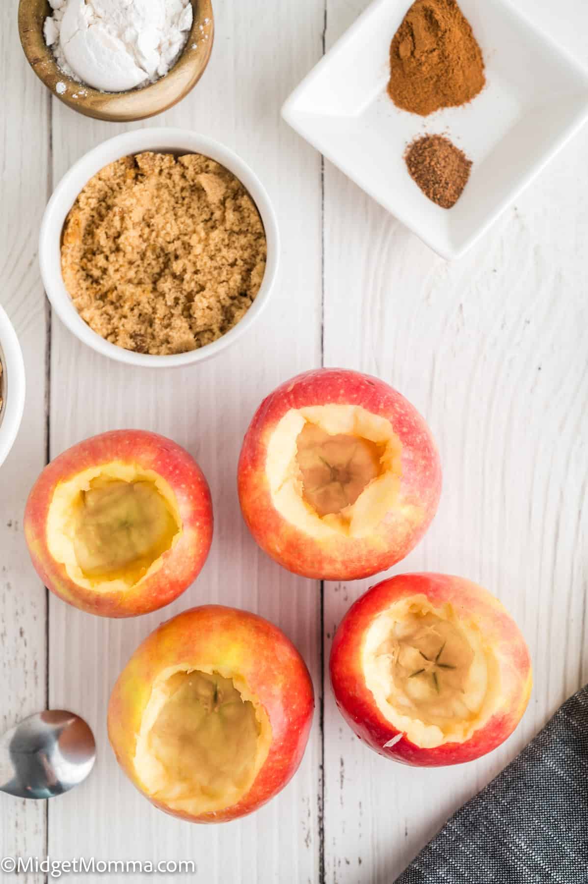 Four cored apples are arranged on a white wooden surface, surrounded by bowls of brown sugar, flour, and spices.