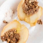 Two baked apple halves on a white plate, each filled with an oat and nut crumble, served with a scoop of vanilla ice cream.