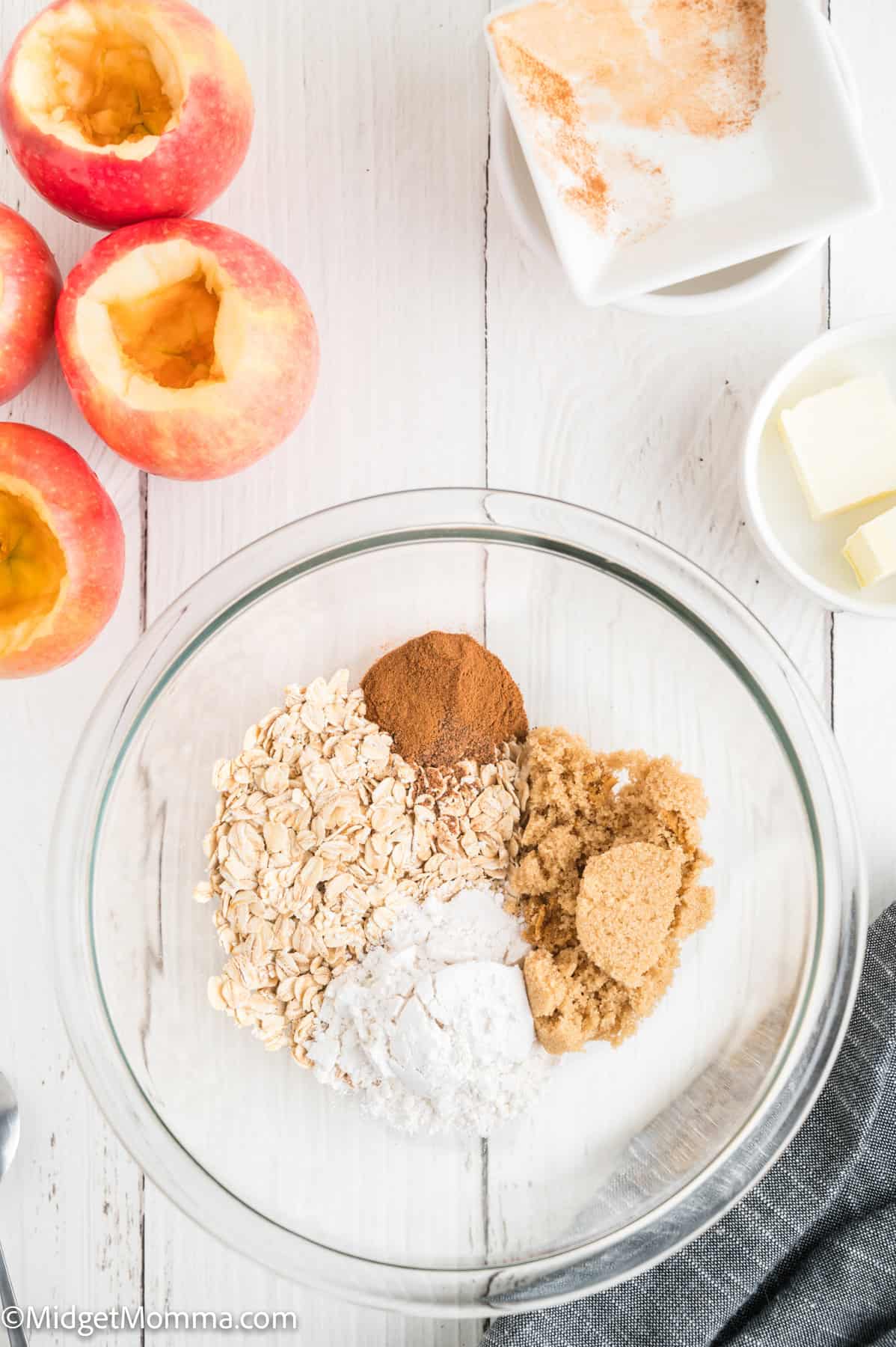 Clear glass bowl with oats, flour, brown sugar, and cinnamon on a white table next to halved apples, a dish of butter, and a small plate with spices.