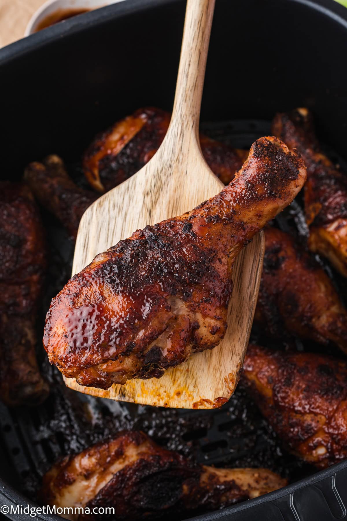 A wooden spatula holds a cooked, seasoned chicken drumstick over a tray of several other drumsticks.