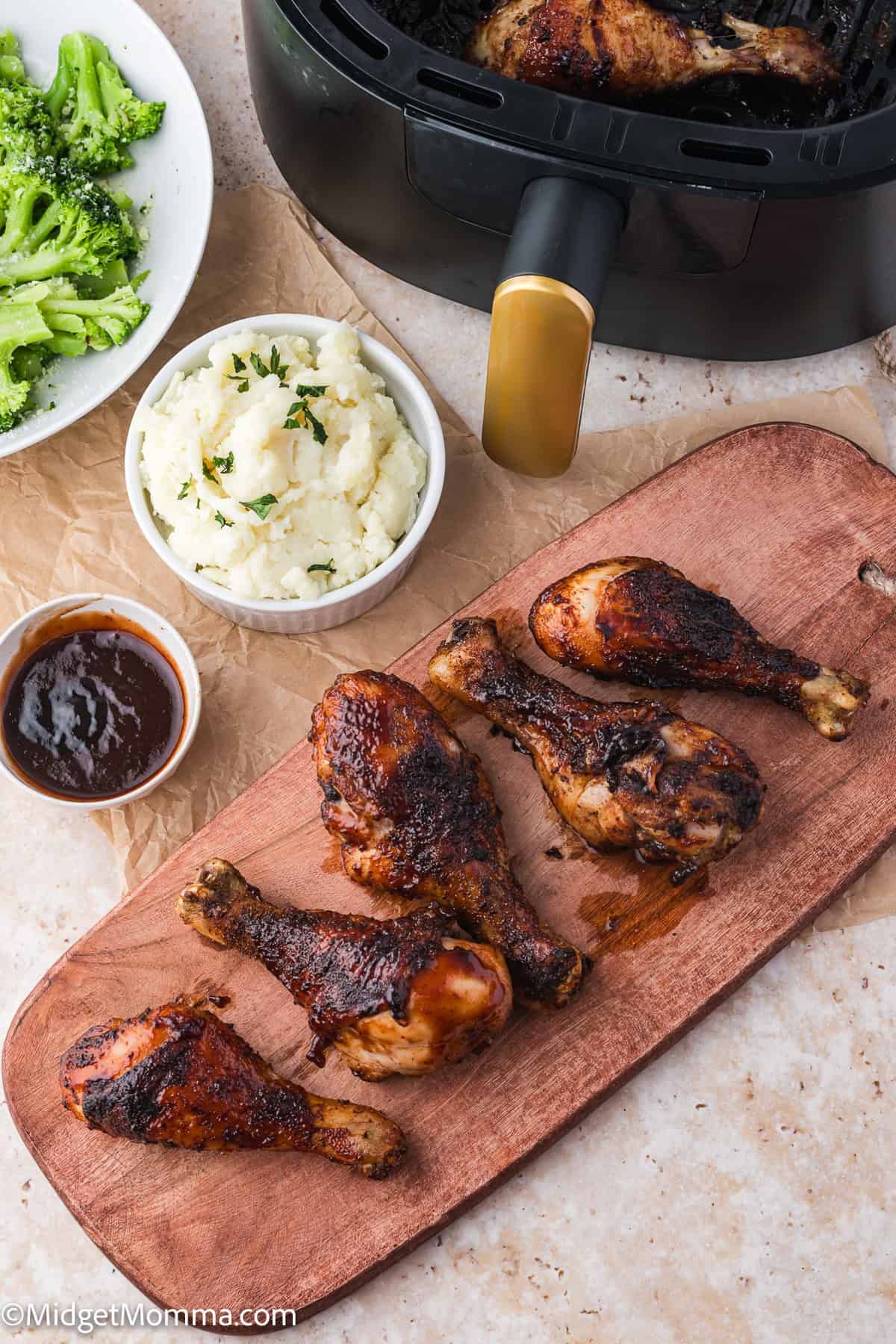 Five cooked chicken drumsticks on a wooden board, with a bowl of mashed potatoes, broccoli, and a small dish of sauce beside an air fryer.