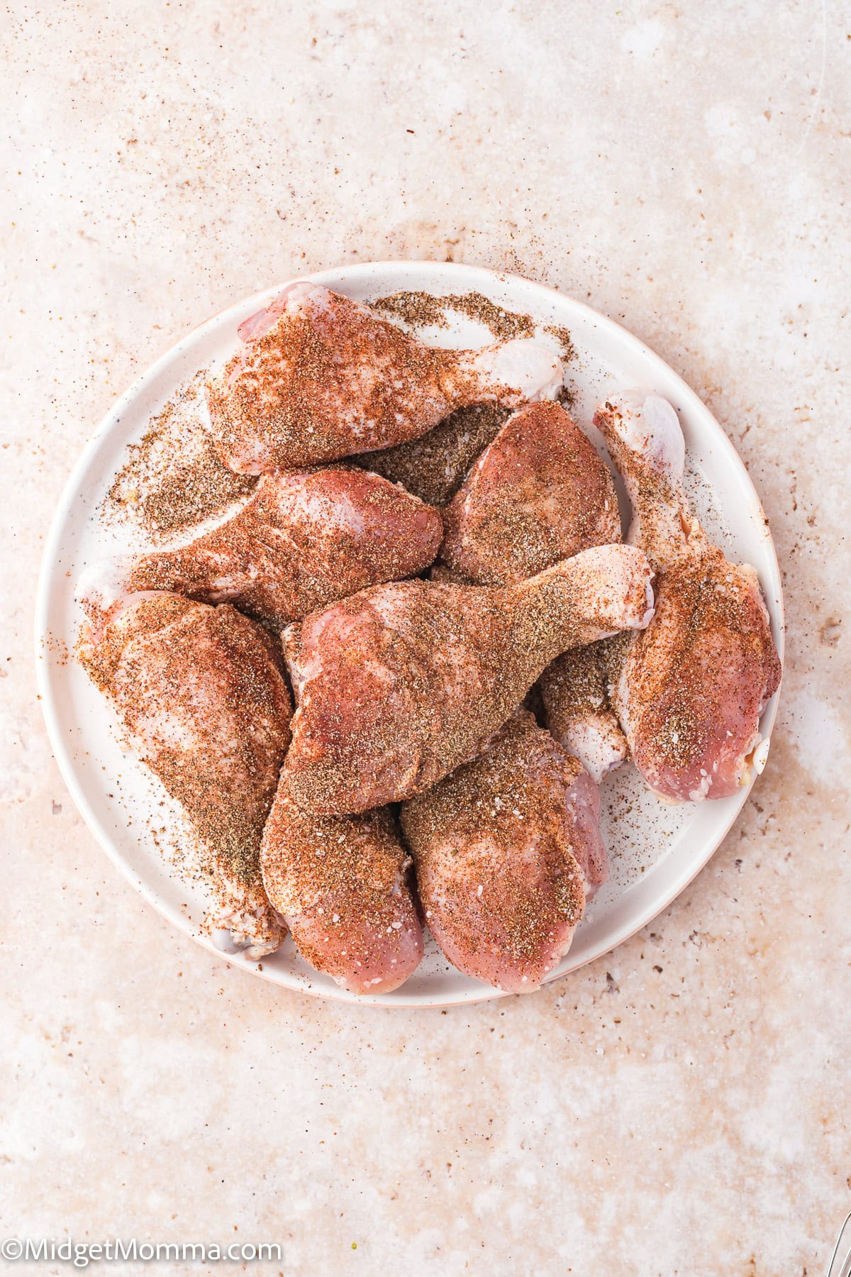 A plate of raw chicken drumsticks coated with a blend of dry seasonings on a light-colored surface.