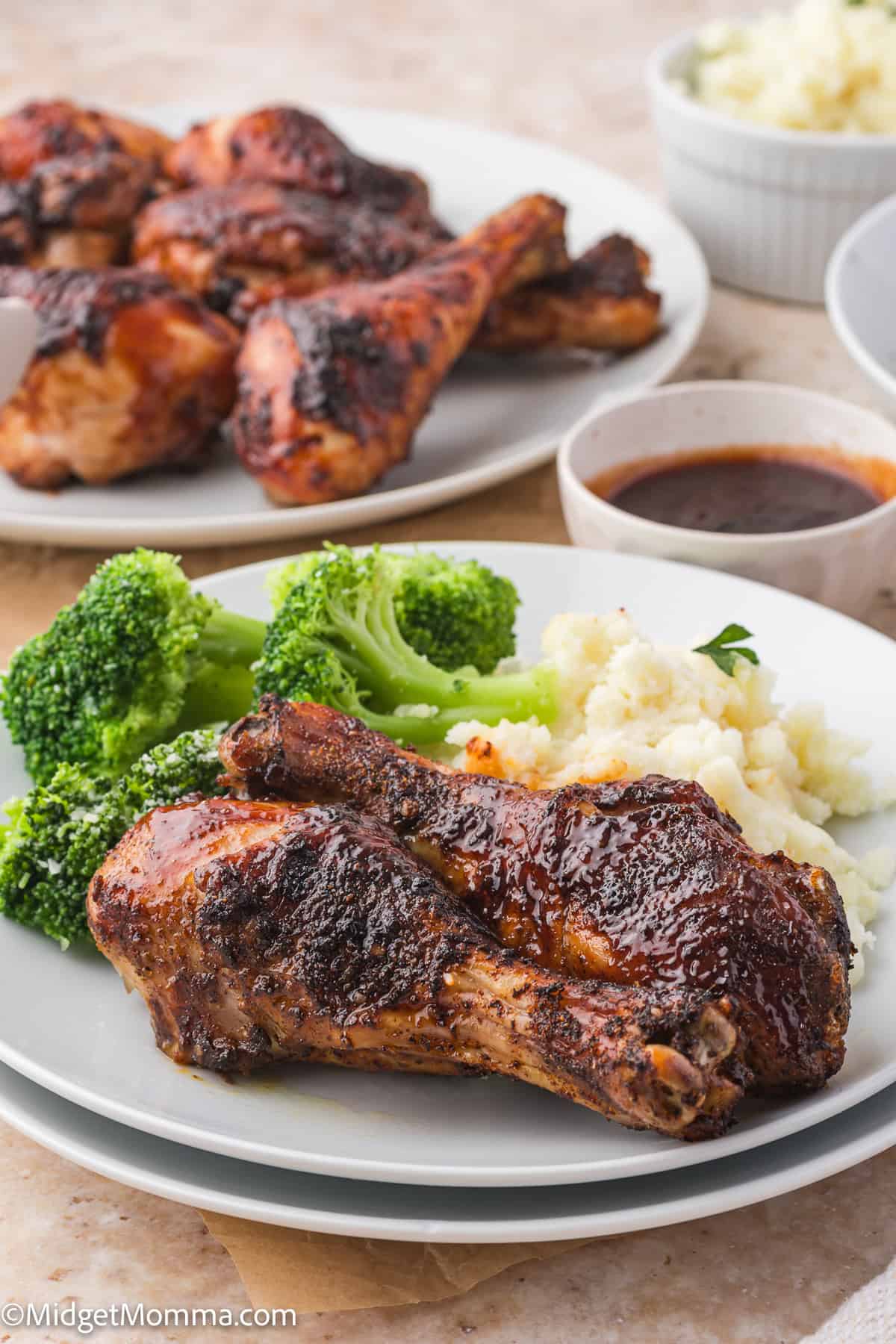 A plate with two grilled chicken drumsticks, steamed broccoli, and mashed potatoes. Another plate of drumsticks and bowls of sauce and mashed potatoes are in the background.