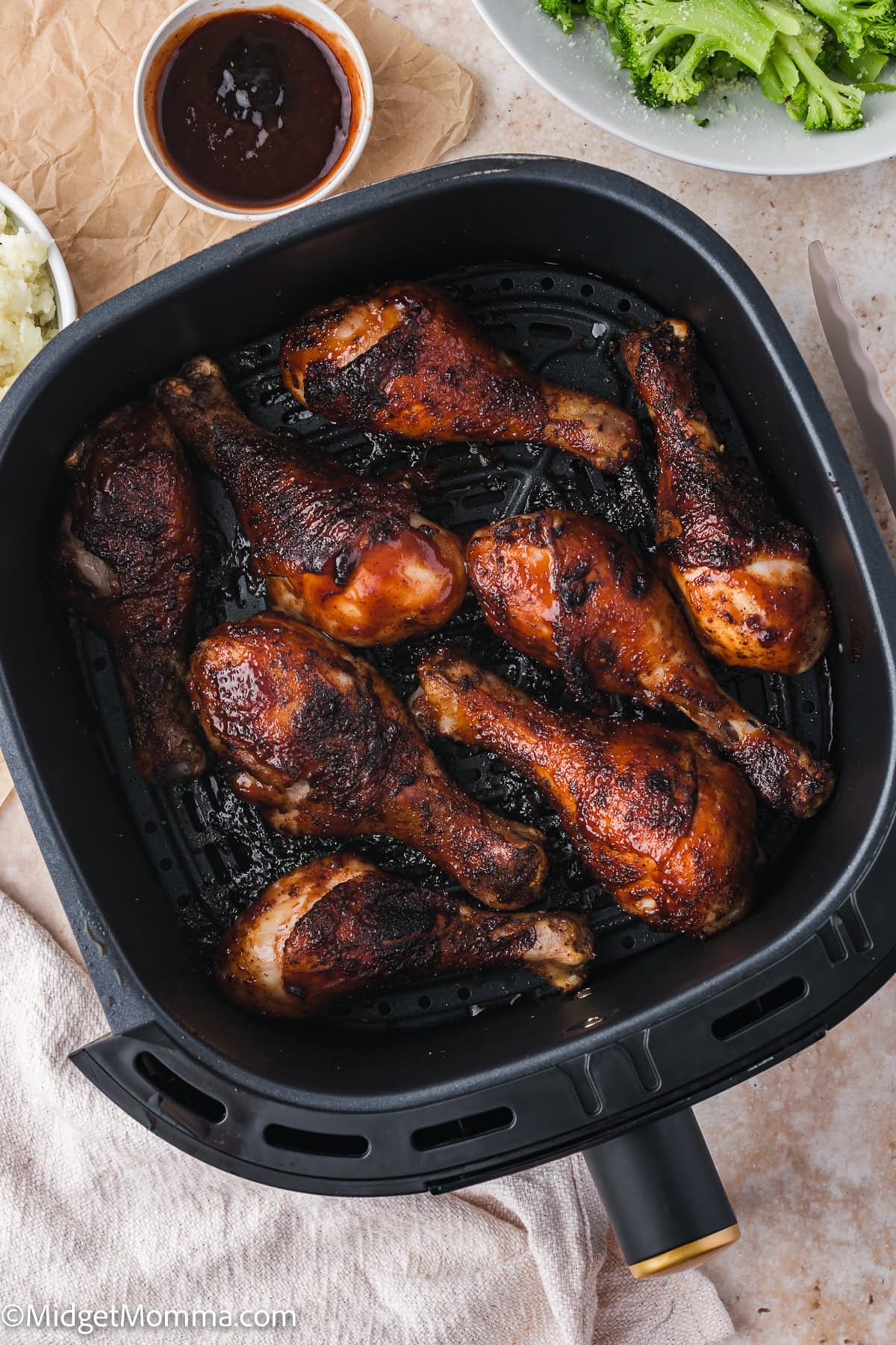 Eight cooked chicken drumsticks in an air fryer basket, with barbecue sauce in a small bowl and broccoli on the side.