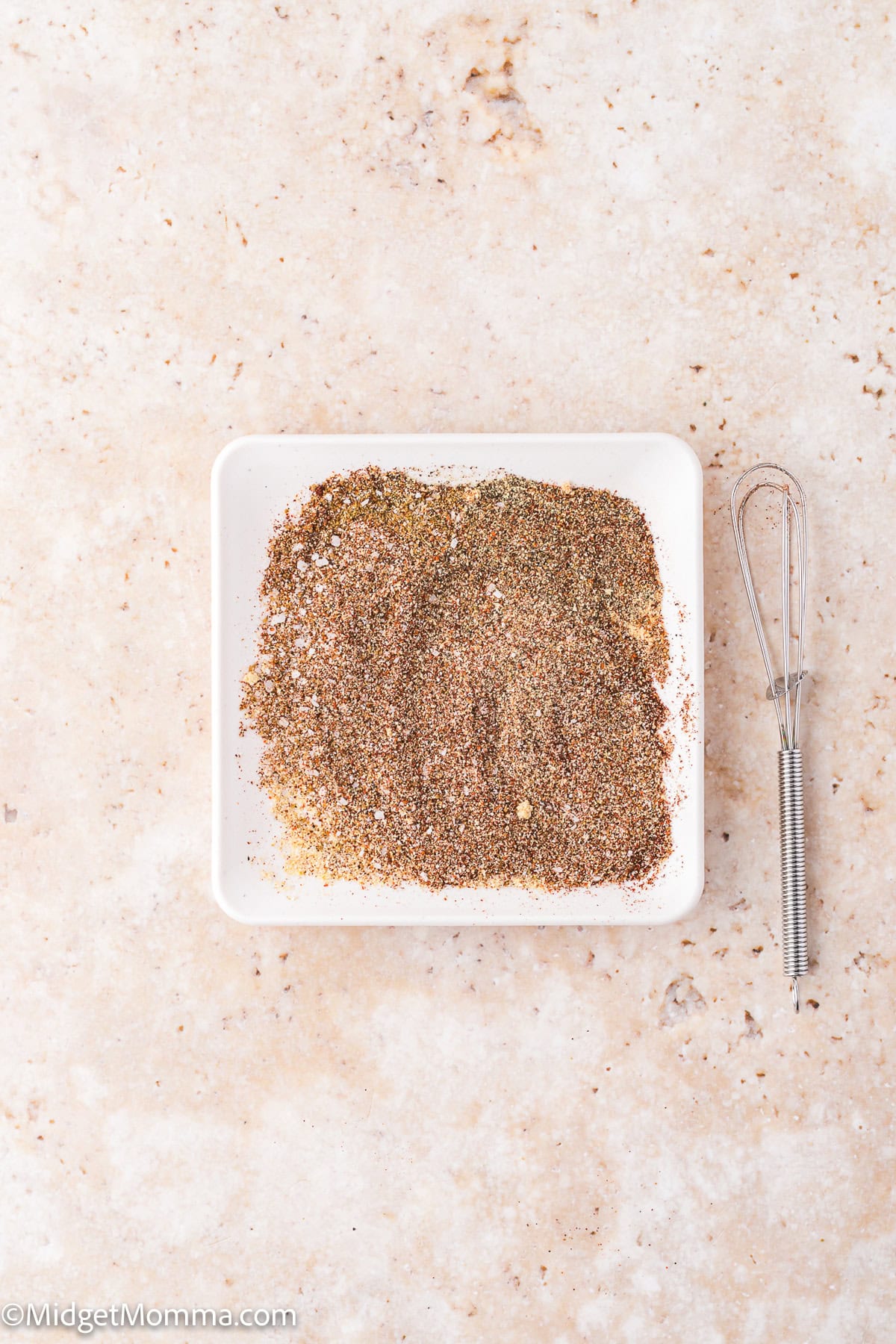 A square white dish filled with a mixed seasoning blend sits on a light countertop next to a small metal whisk.