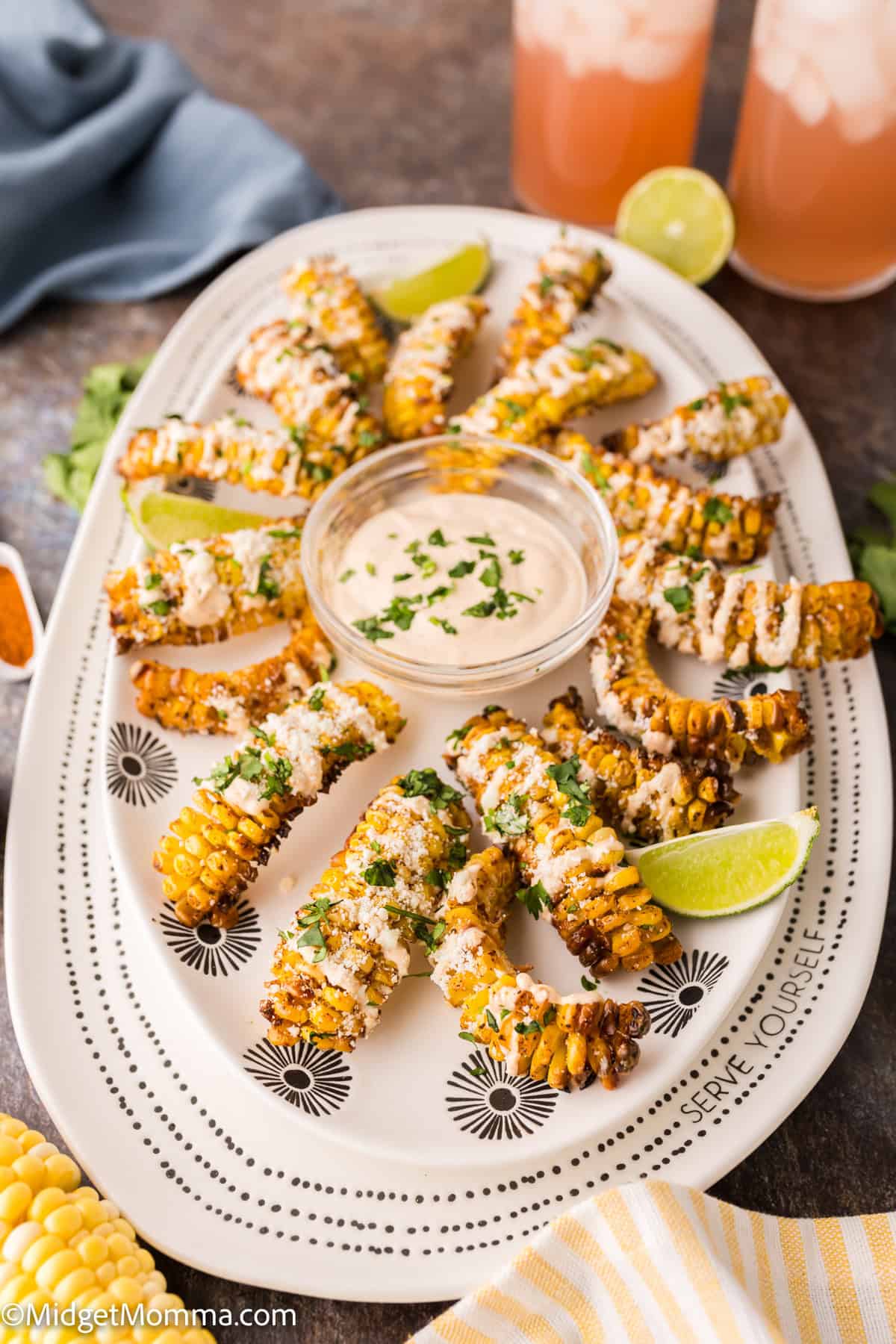 A platter of grilled corn ribs garnished with cheese and herbs, arranged around a bowl of dipping sauce, with lime wedges and two iced drinks in the background.