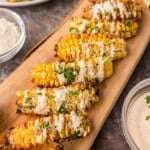 Grilled corn ribs arranged on a wooden board, topped with a creamy sauce and chopped herbs, with bowls of sauce and grated cheese nearby.
