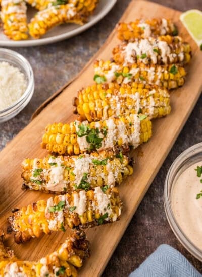 Grilled corn ribs arranged on a wooden board, topped with a creamy sauce and chopped herbs, with bowls of sauce and grated cheese nearby.
