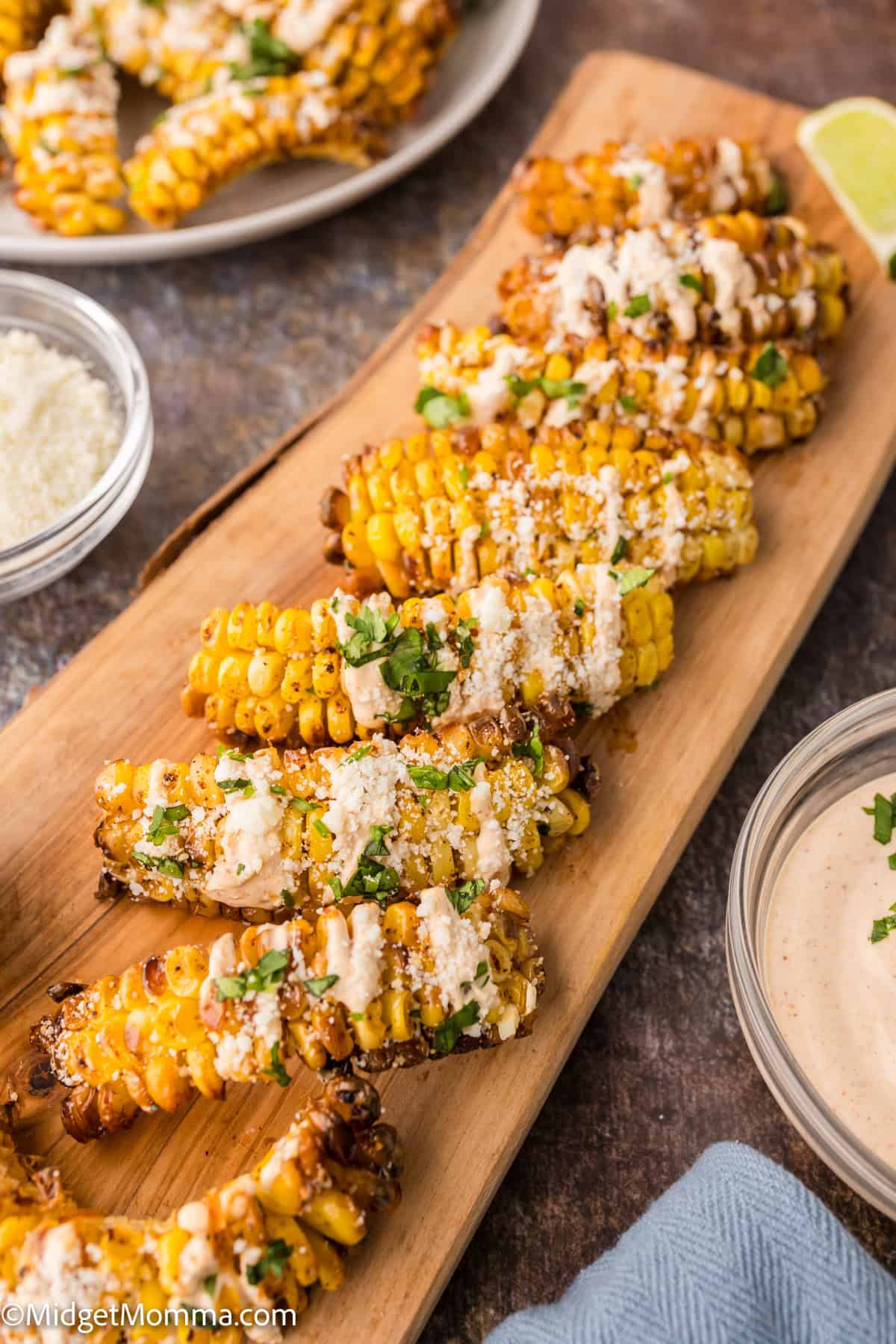 Grilled corn ribs arranged on a wooden board, topped with a creamy sauce and chopped herbs, with bowls of sauce and grated cheese nearby.