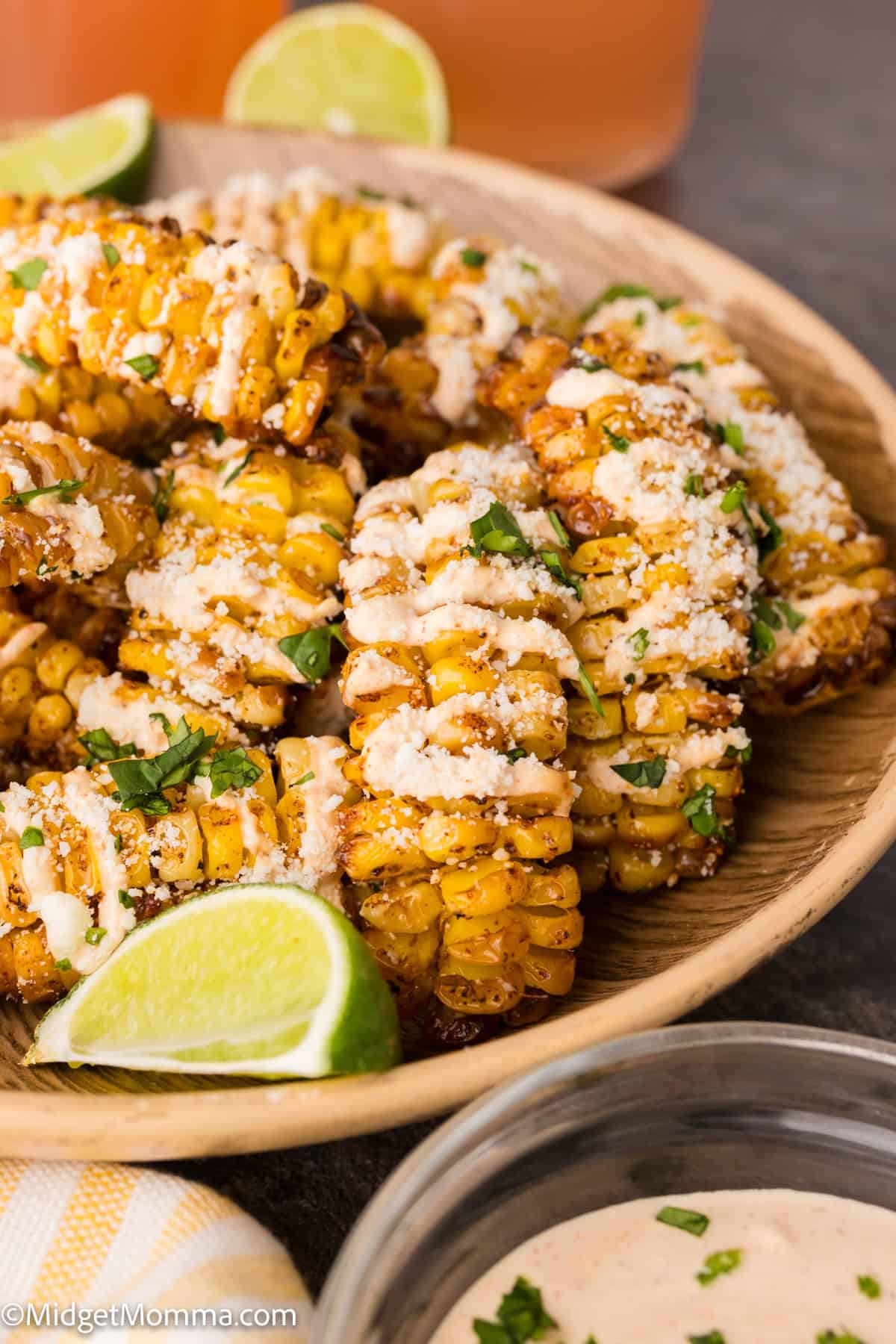 A wooden bowl filled with grilled corn ribs topped with cheese, chopped herbs, and sauce, garnished with lime wedges.