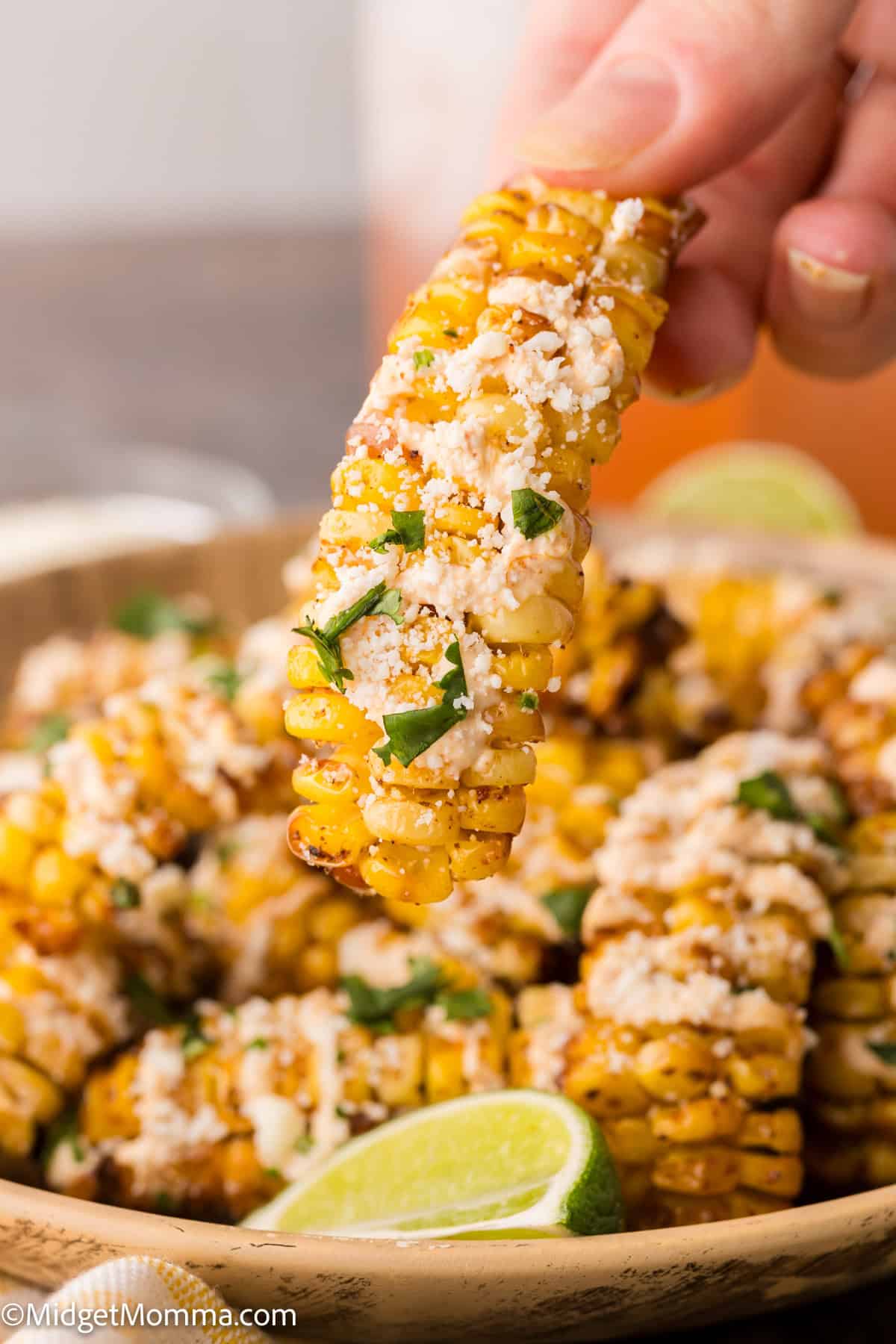 A hand holds a piece of grilled corn rib topped with cheese and herbs, with more corn pieces and a lime wedge on a plate.