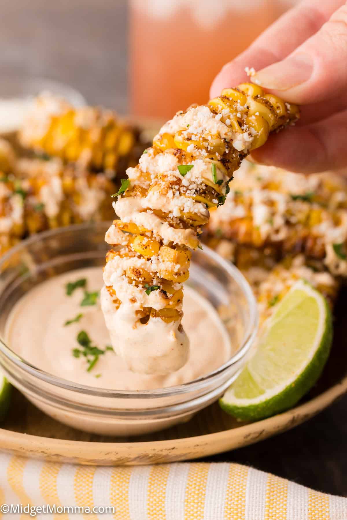 A hand dips a piece of seasoned corn rib into a bowl of creamy sauce, with lime wedges on the plate.