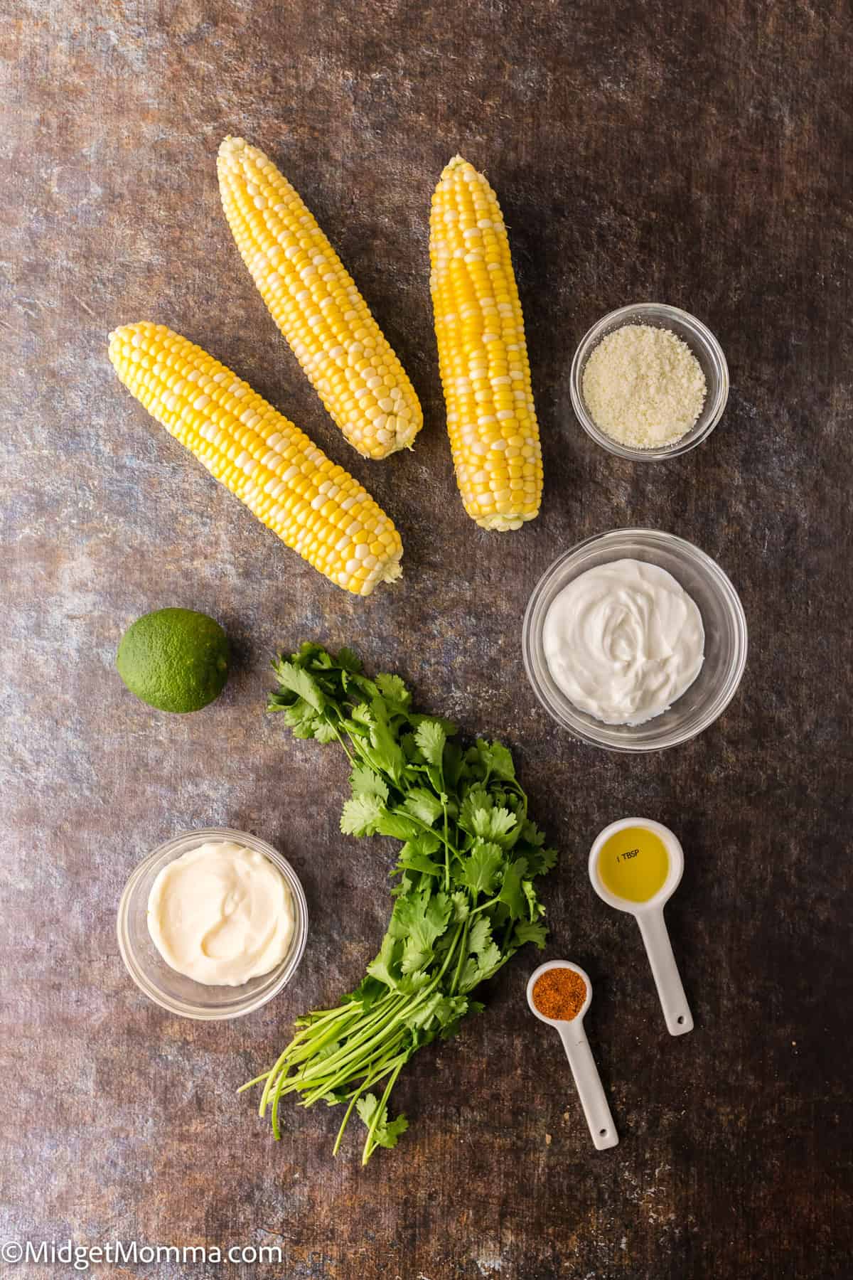 Three ears of corn, a bunch of cilantro, a lime, small bowls of sauce, cheese, and spices arranged on a dark surface.