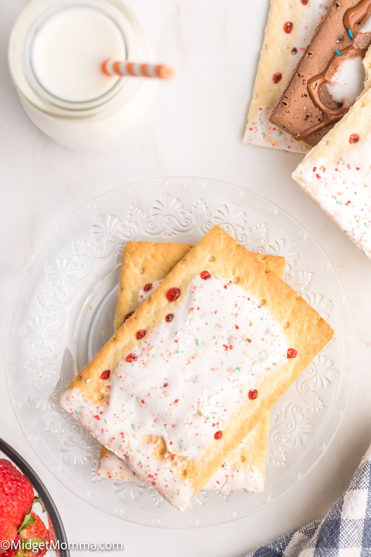 Two frosted rectangular pastries with sprinkles on a glass plate, next to a bottle of milk with a straw and a chocolate frosted pastry on a white surface.