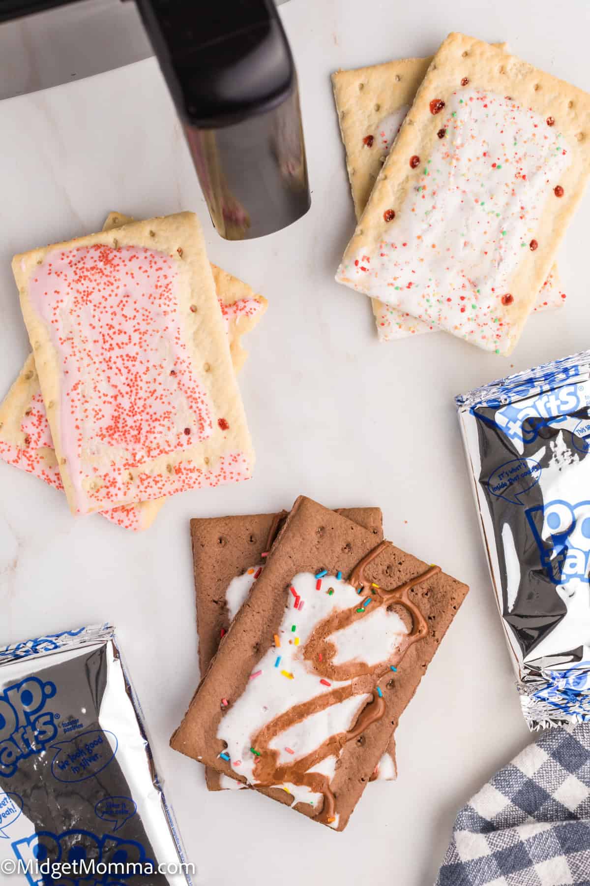 Three types of Pop-Tarts, some opened and some still in silver packaging, are arranged on a white surface near a black appliance.