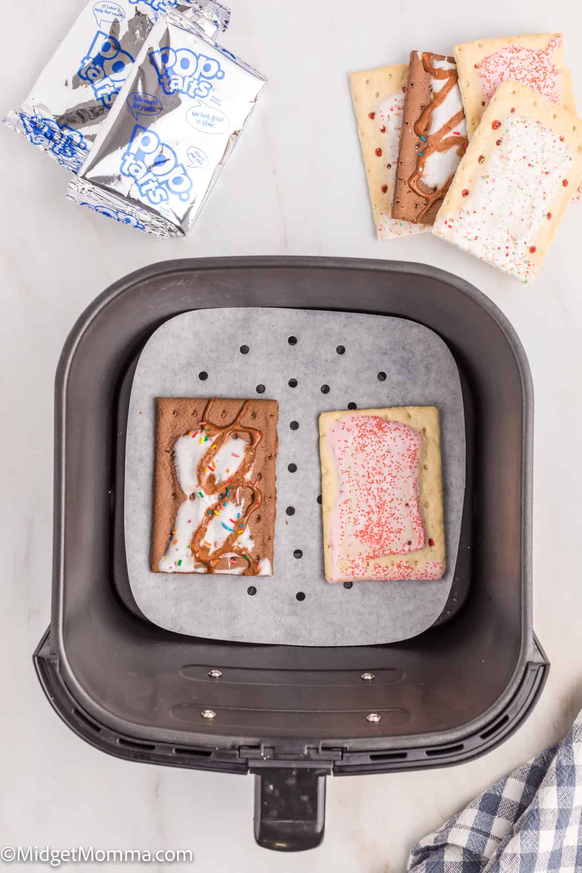 Two Pop-Tarts, one chocolate and one strawberry, are placed on parchment paper inside an air fryer basket, with additional Pop-Tarts and wrappers nearby.