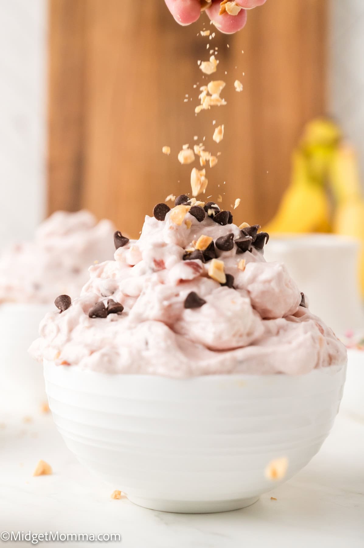 A white bowl filled with pink creamy dessert topped with chocolate chips, as chopped nuts are sprinkled on top. Bananas and another bowl are blurred in the background.