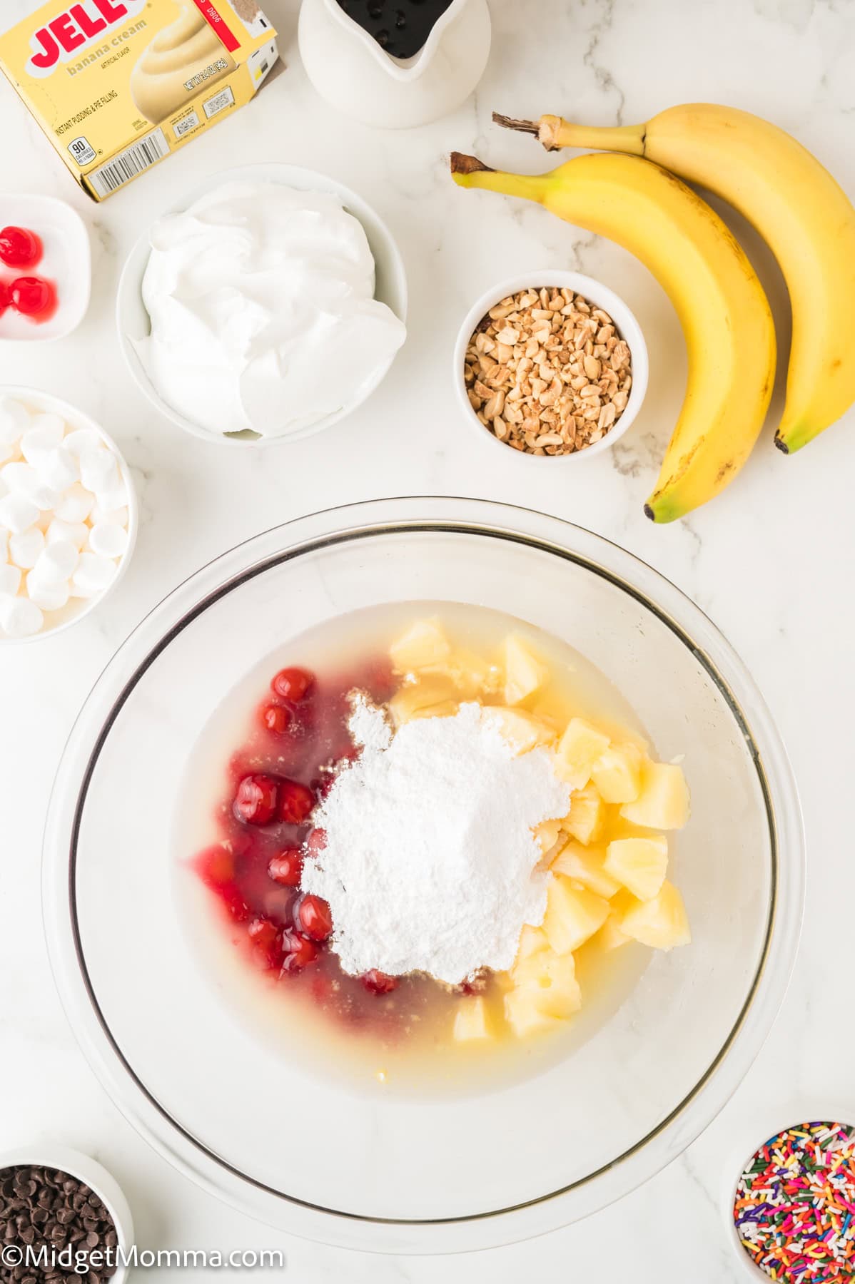 A glass bowl with pineapple chunks, cherries, and powdered sugar, surrounded by bananas, whipped topping, marshmallows, sprinkles, nuts, and a box of Jell-O pudding mix on a countertop.